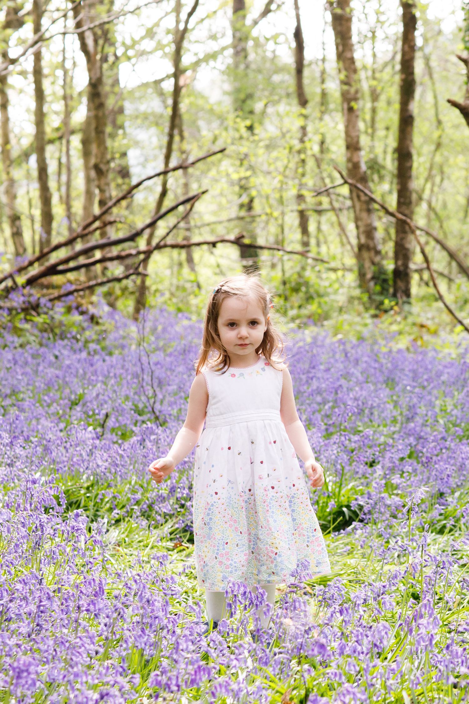 Child in white dress standing among purple bluebells in spring woodland.