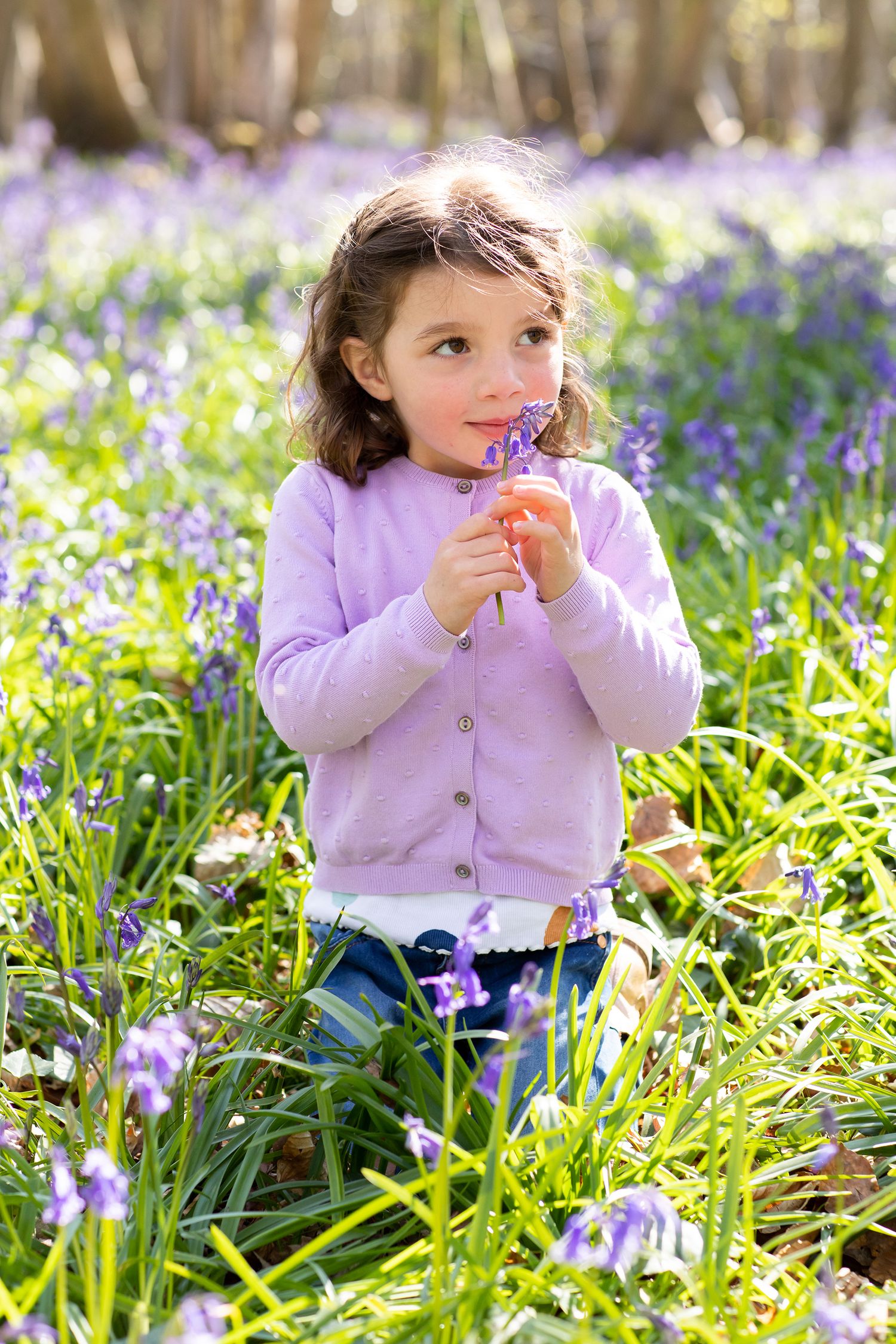 Young child in purple cardigan smelling bluebells in a spring woodland.