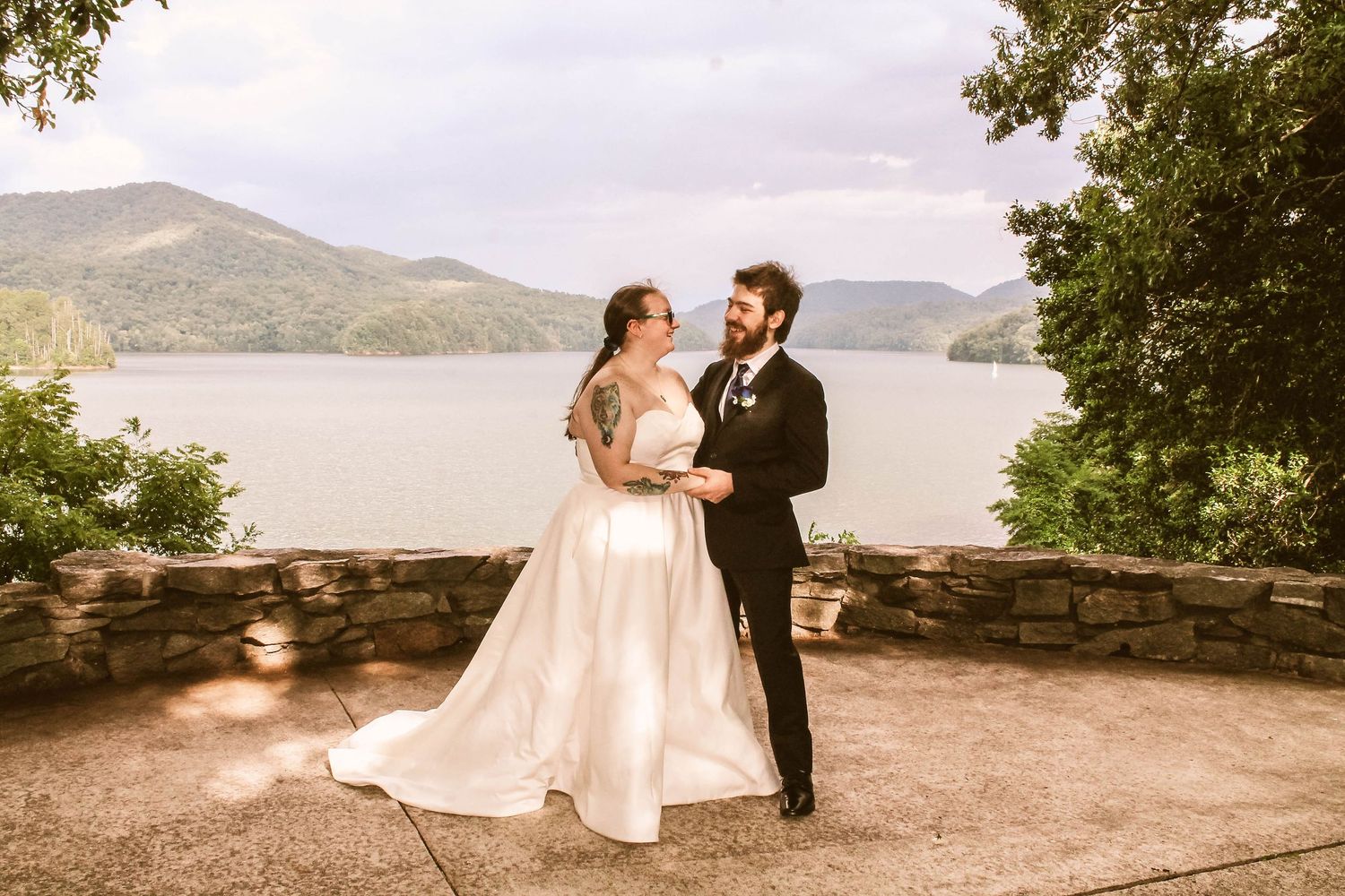 A couple in wedding attire poses on a scenic overlook with mountains and water in the background.