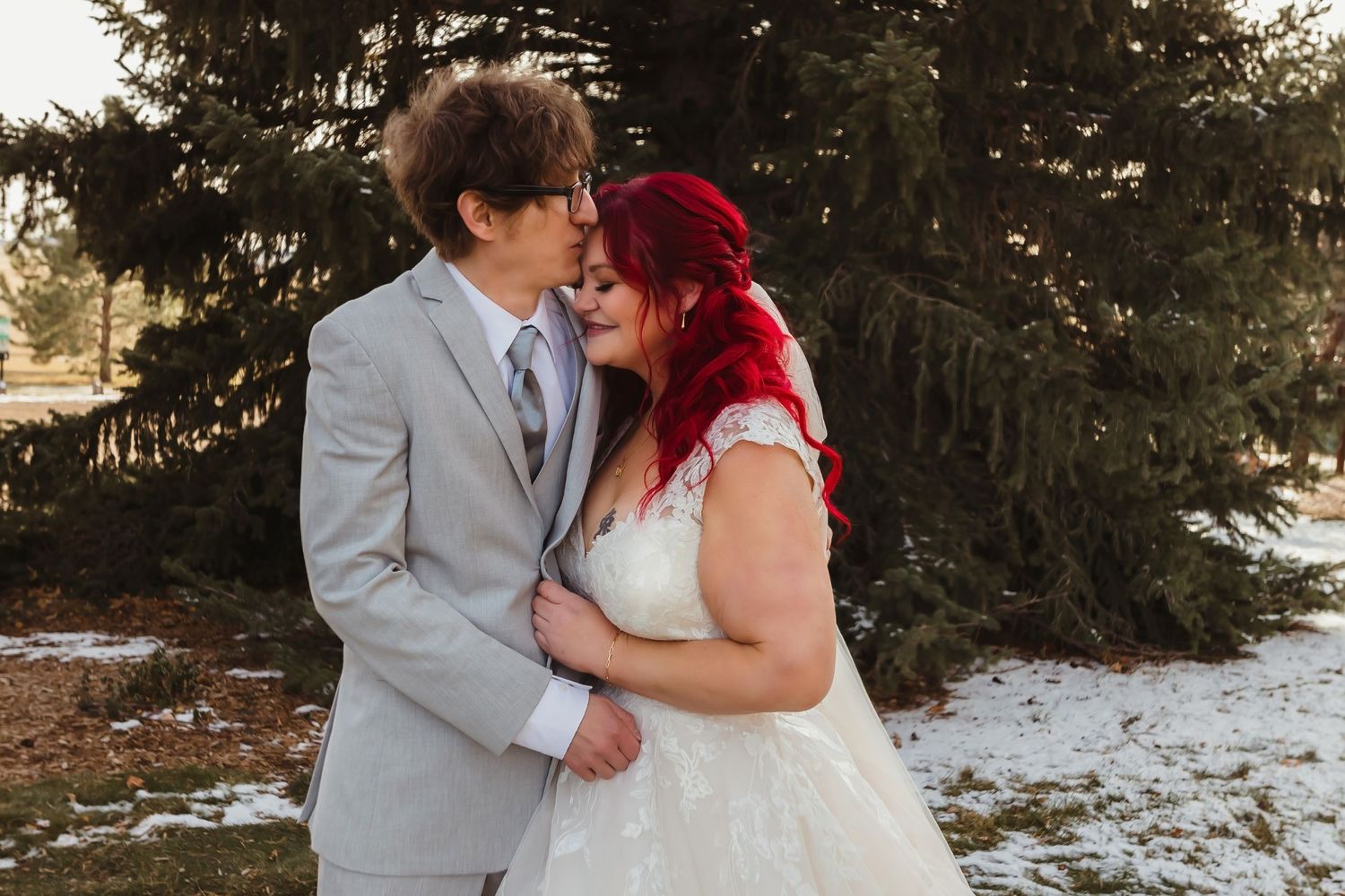 A winter wedding portrait featuring newlyweds embracing in front of evergreen trees and snow.
