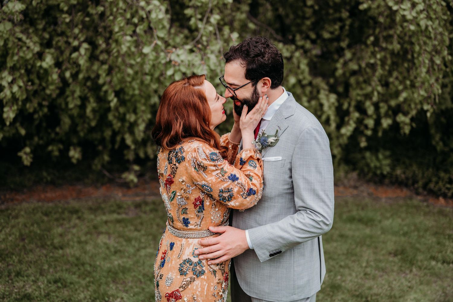 A couple in a romantic embrace outdoors, with a woman in floral dress and man in grey blazer standing on grass in the Hudson Valley.