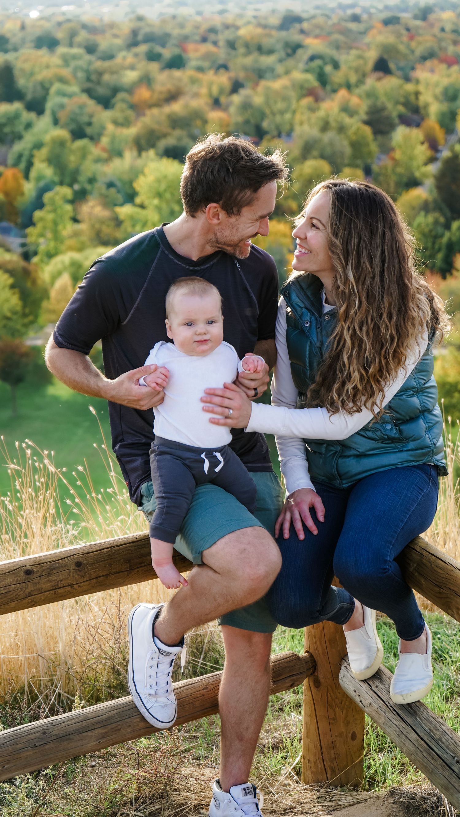 A family sits together on a wooden fence overlooking an autumn landscape with colorful foliage.