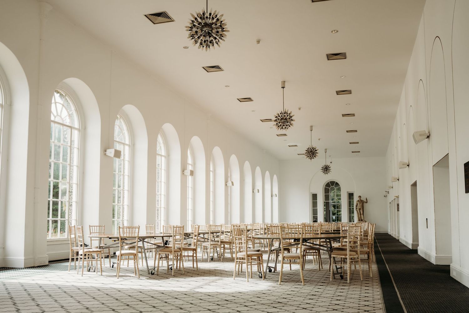 Bright sunlit hall with arched windows and wooden chairs arranged for an event.