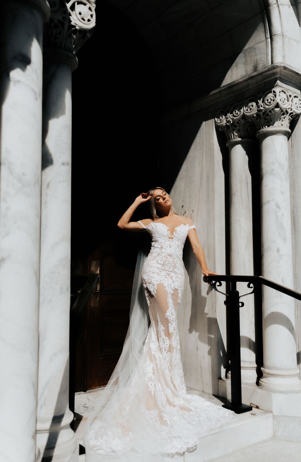 Bride in lace see-through wedding gown poses dramatically on historic building steps.