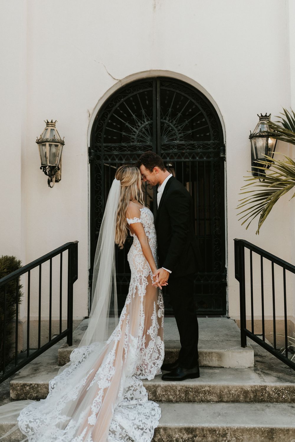 Romantic wedding couple stand in front of large arched doorway with ornate lanterns on either side.