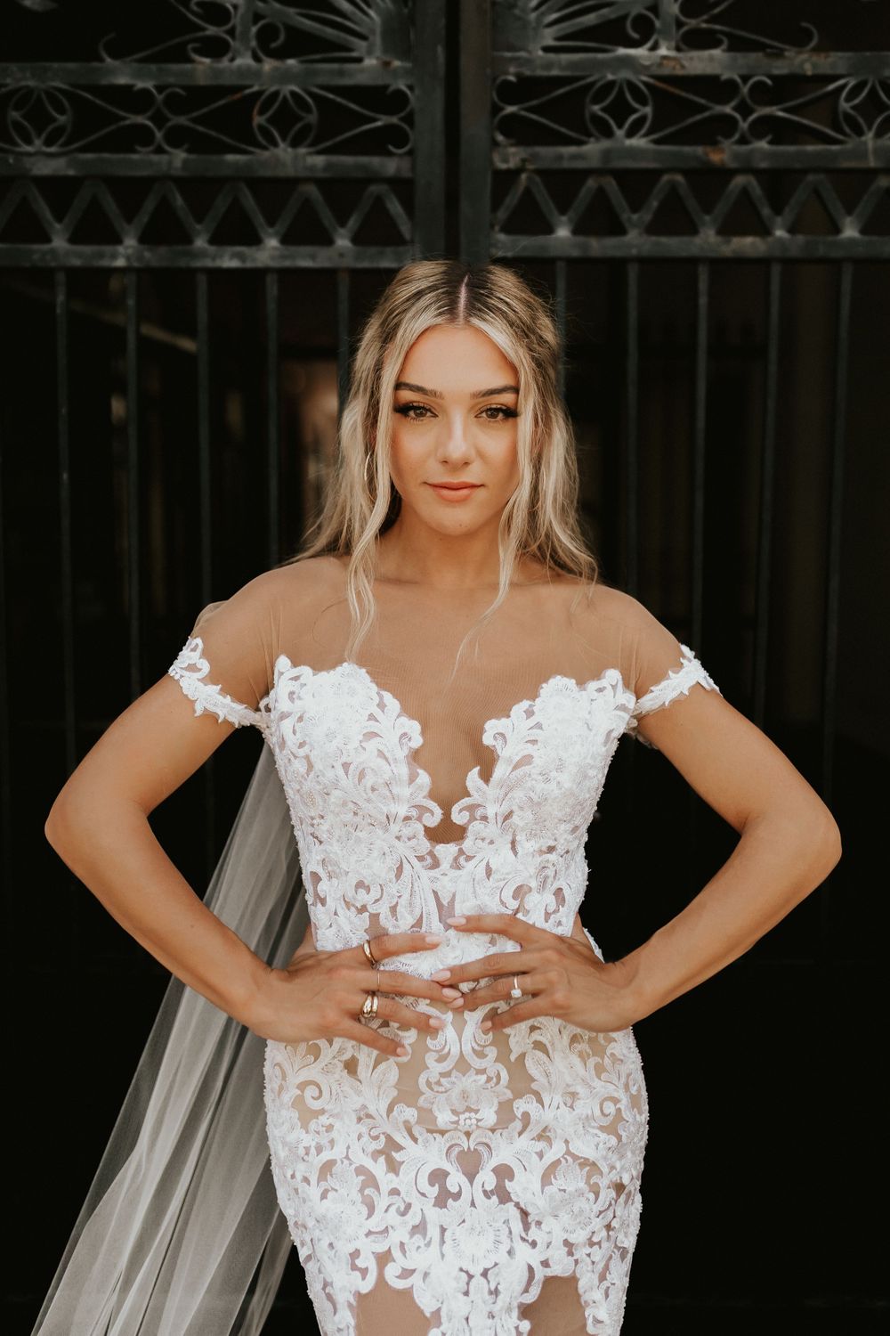 Bride in detailed lace wedding dress poses against ornate iron gate.