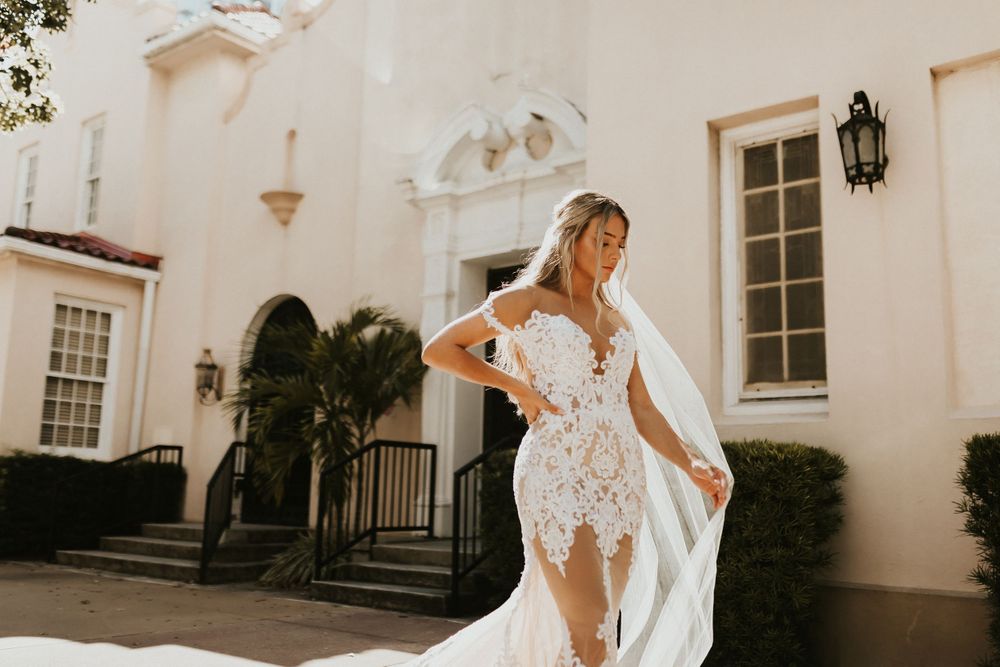 Elegant bride in flowing lace gown poses outside historic building with palm trees.
