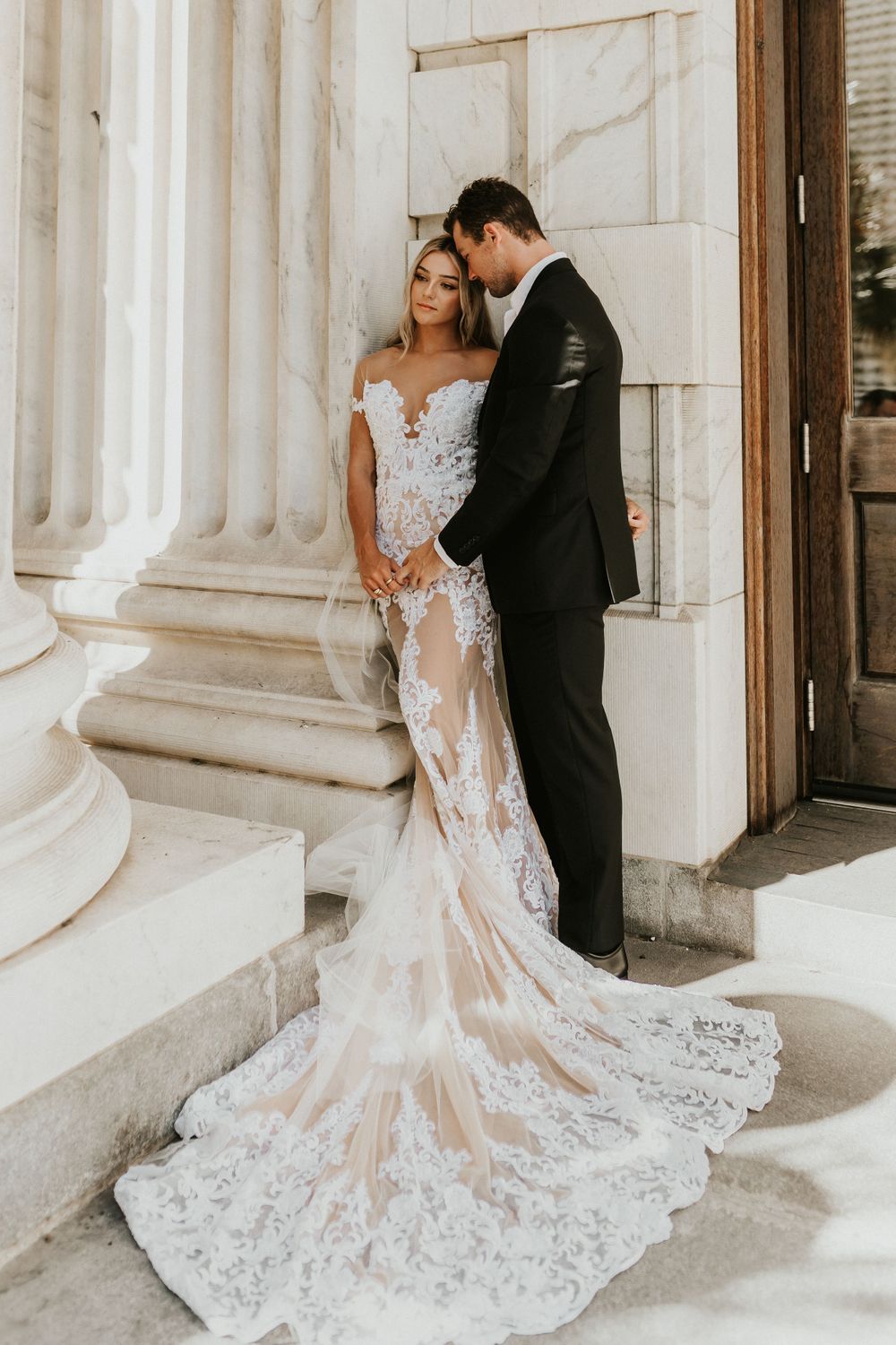 Elegant couple embraces on marble steps with bride's lace dress trailing beautifully.