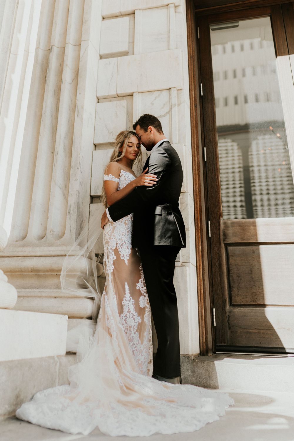 Romantic wedding moment captured against white columned building with flowing lace dress.