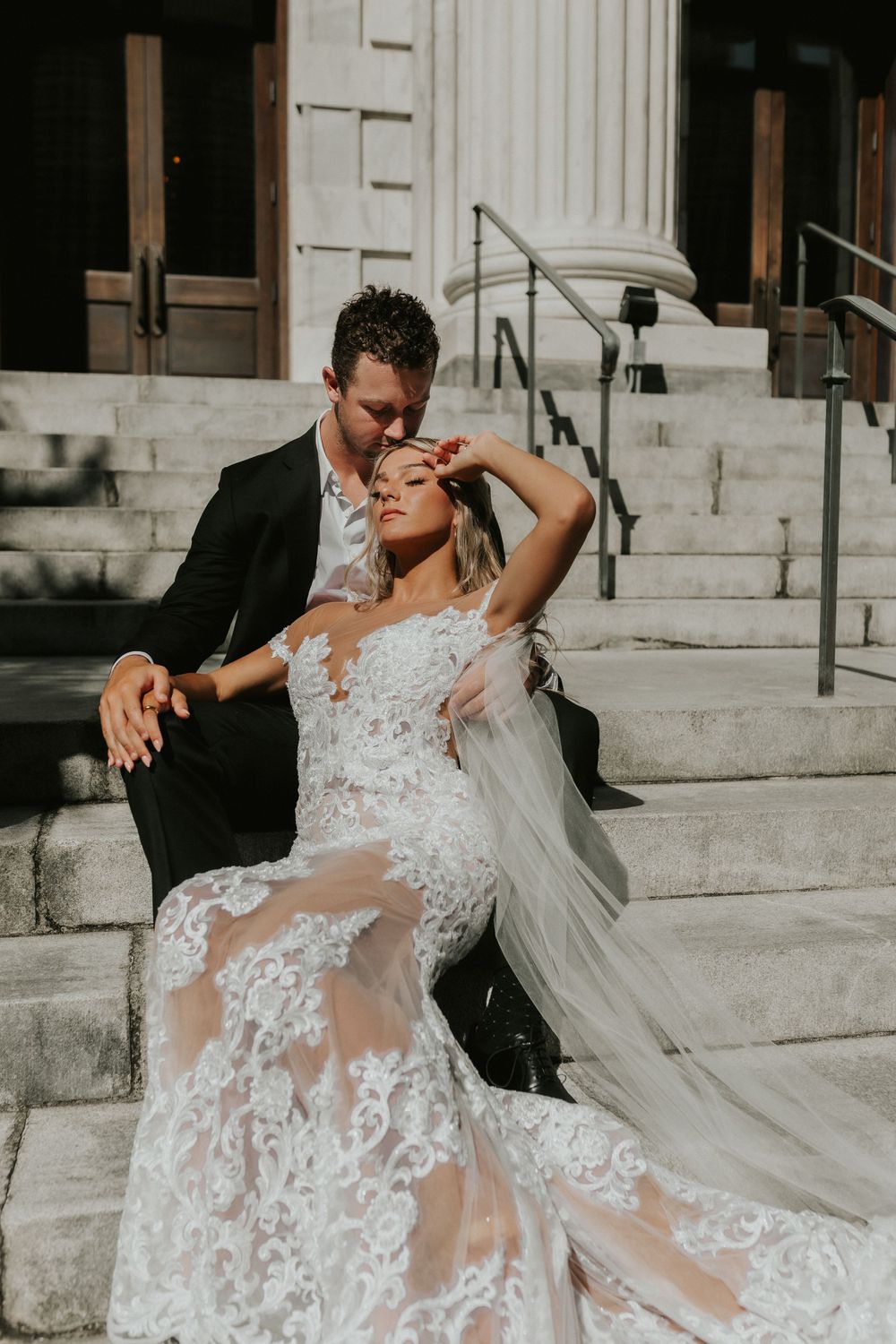 Stylish wedding couple poses romantically on marble steps in warm sunlight.