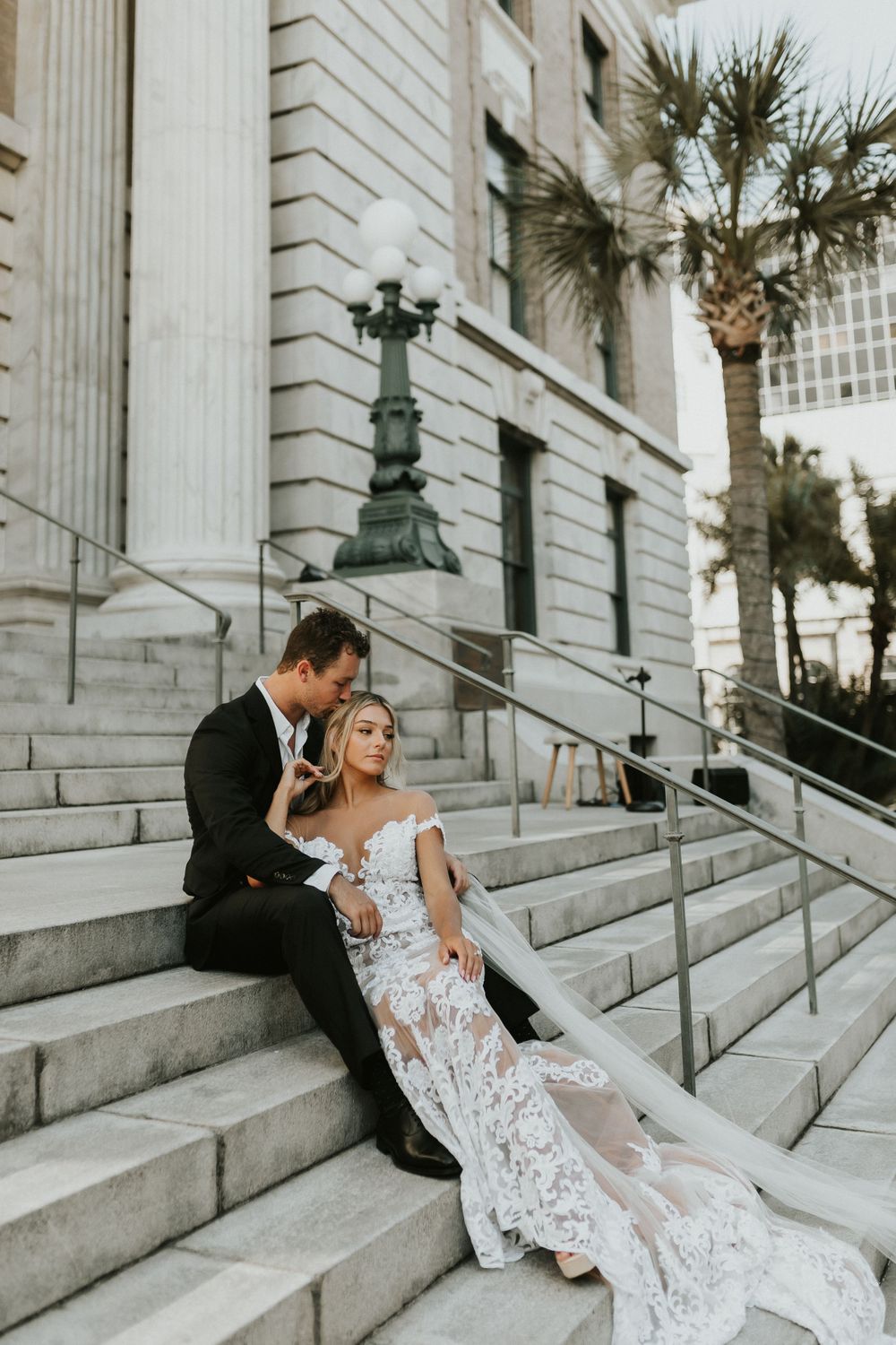 A couple sits on grand stone steps of a historic building with white columns and palm trees in an elegant wedding photo.