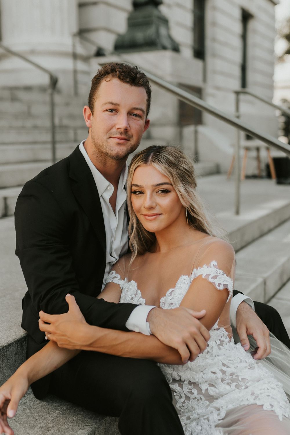A bride in lace gown and groom in black suit pose together on stone steps of a classic building.