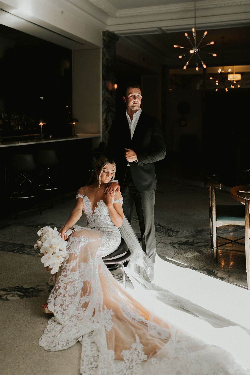 A dramatic evening portrait of a couple, with the bride in a flowing lace gown against moody interior lighting.