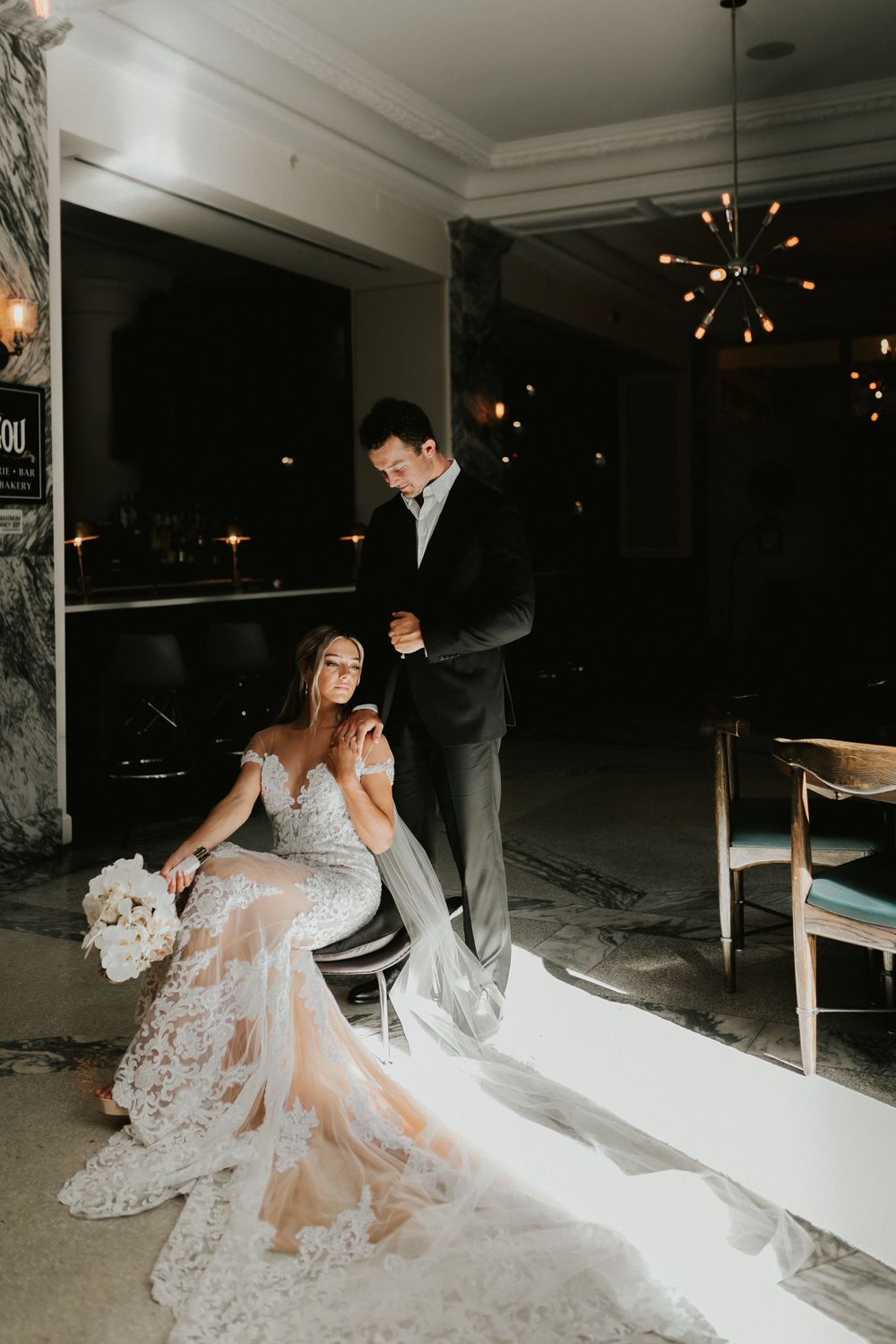 An elegant evening wedding photo with a couple posing near a modern chandelier in a darkened room.