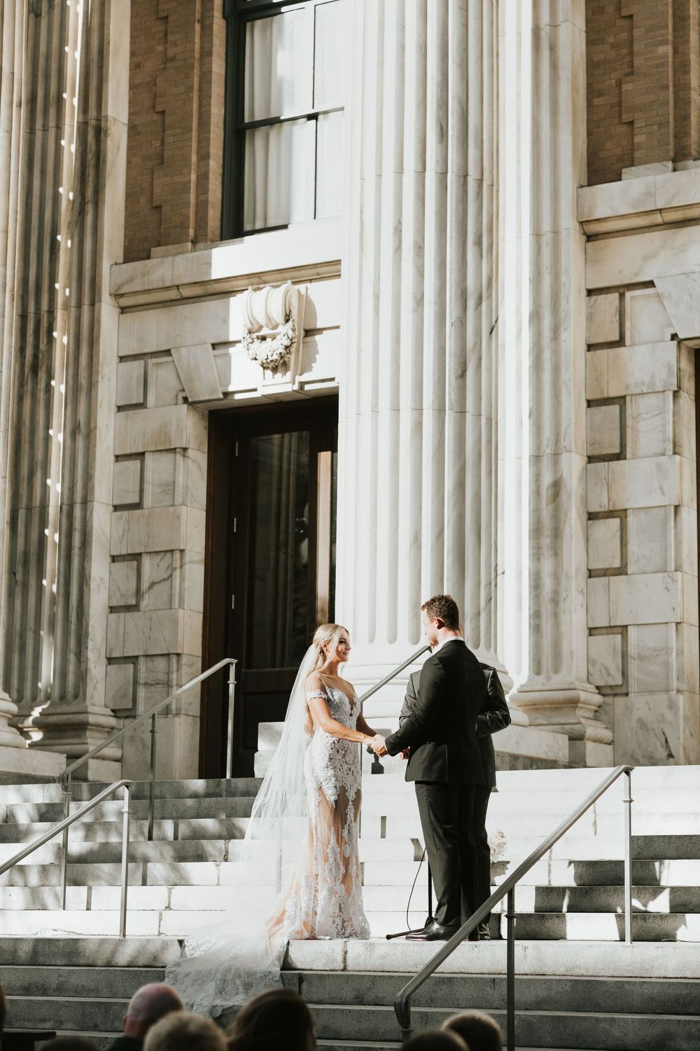 A couple stands on marble steps between classic white columns during their wedding ceremony.