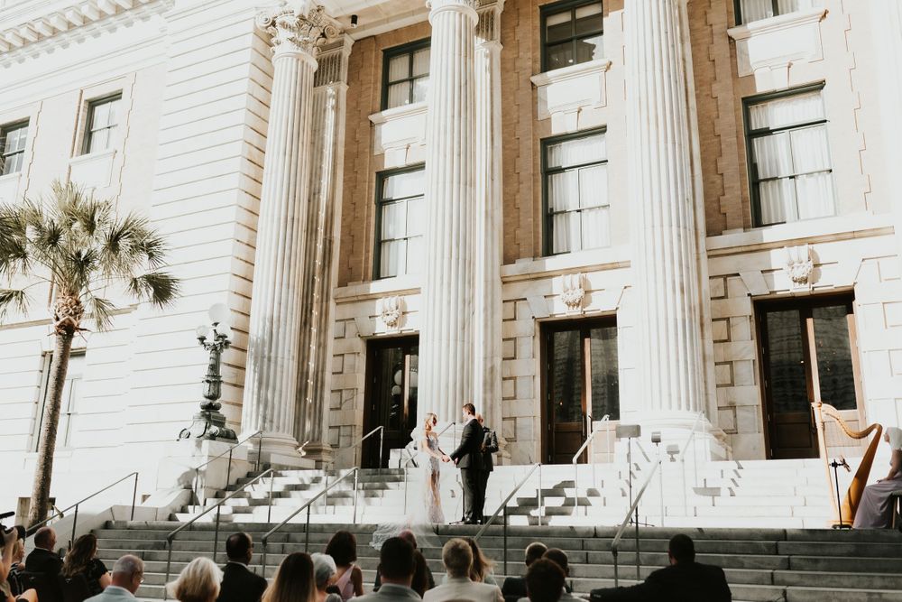 A wedding ceremony takes place on the steps of a grand historic building with white columns and palm trees.