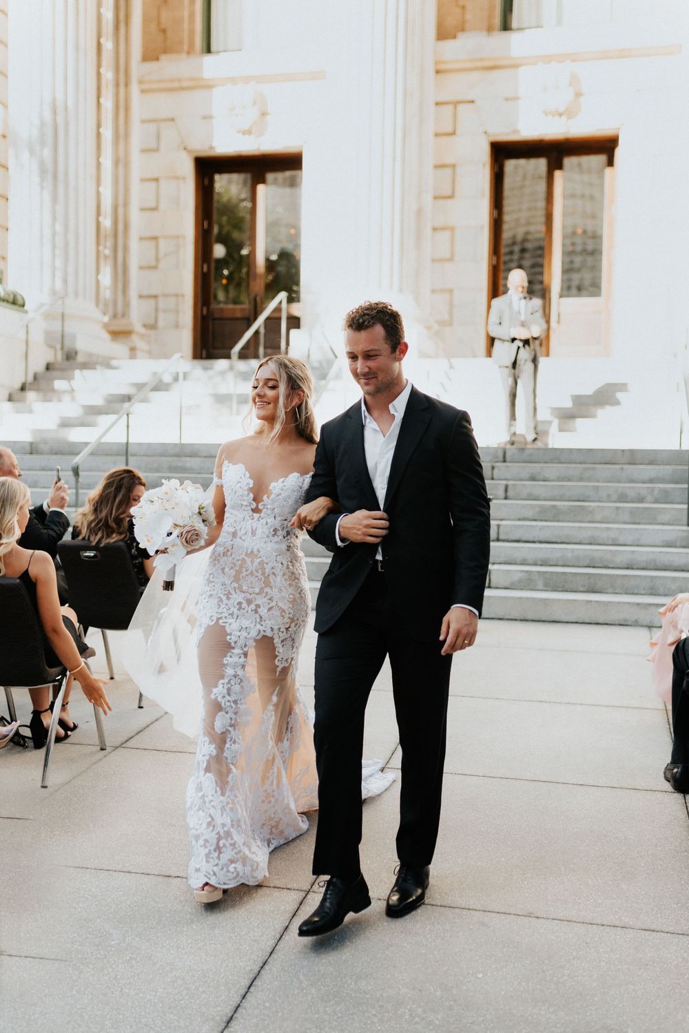 A couple walks past wedding guests on a bright afternoon outside a columned building.