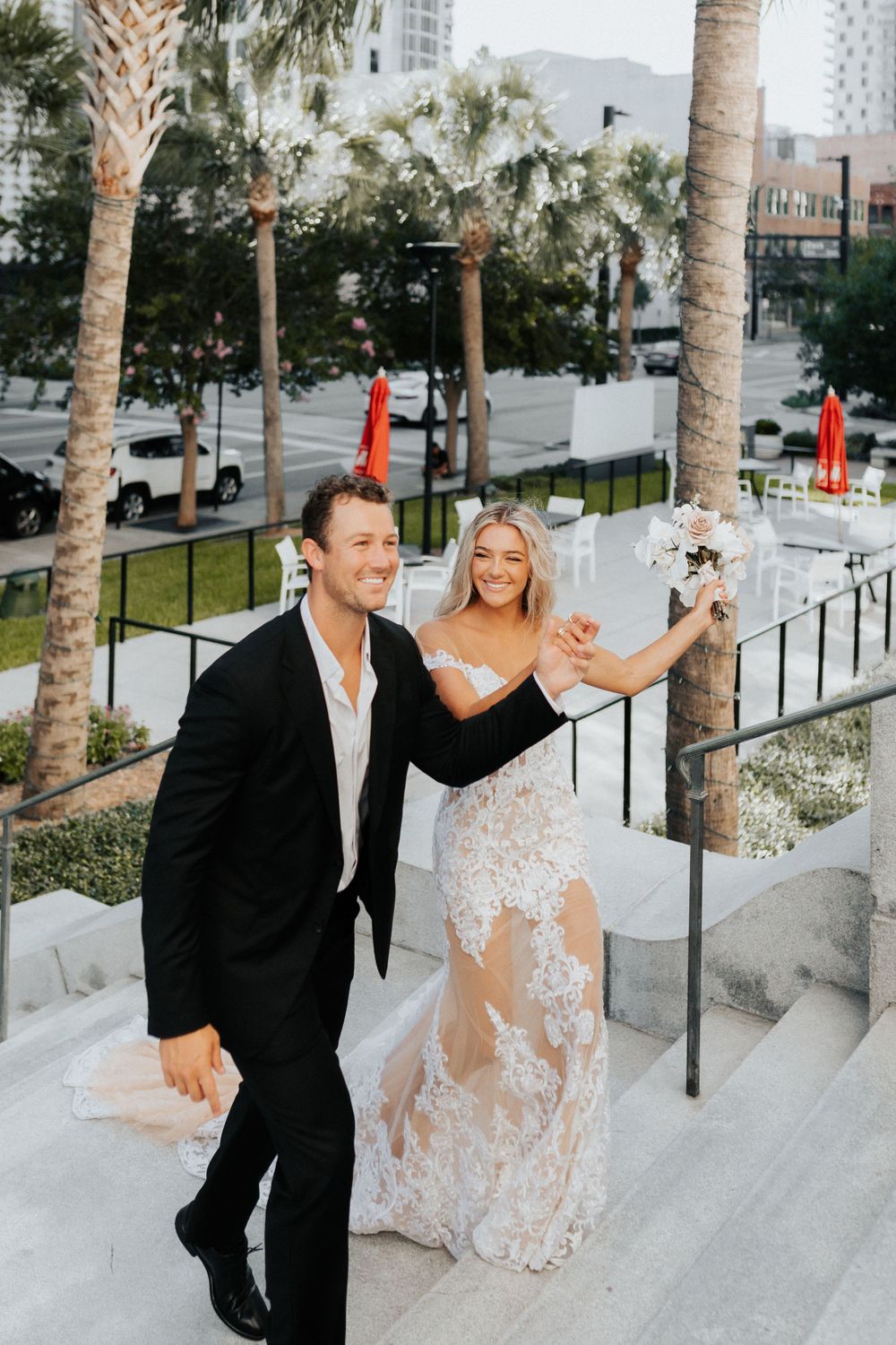 A joyful couple celebrates on outdoor steps surrounded by palm trees in formal wedding attire.