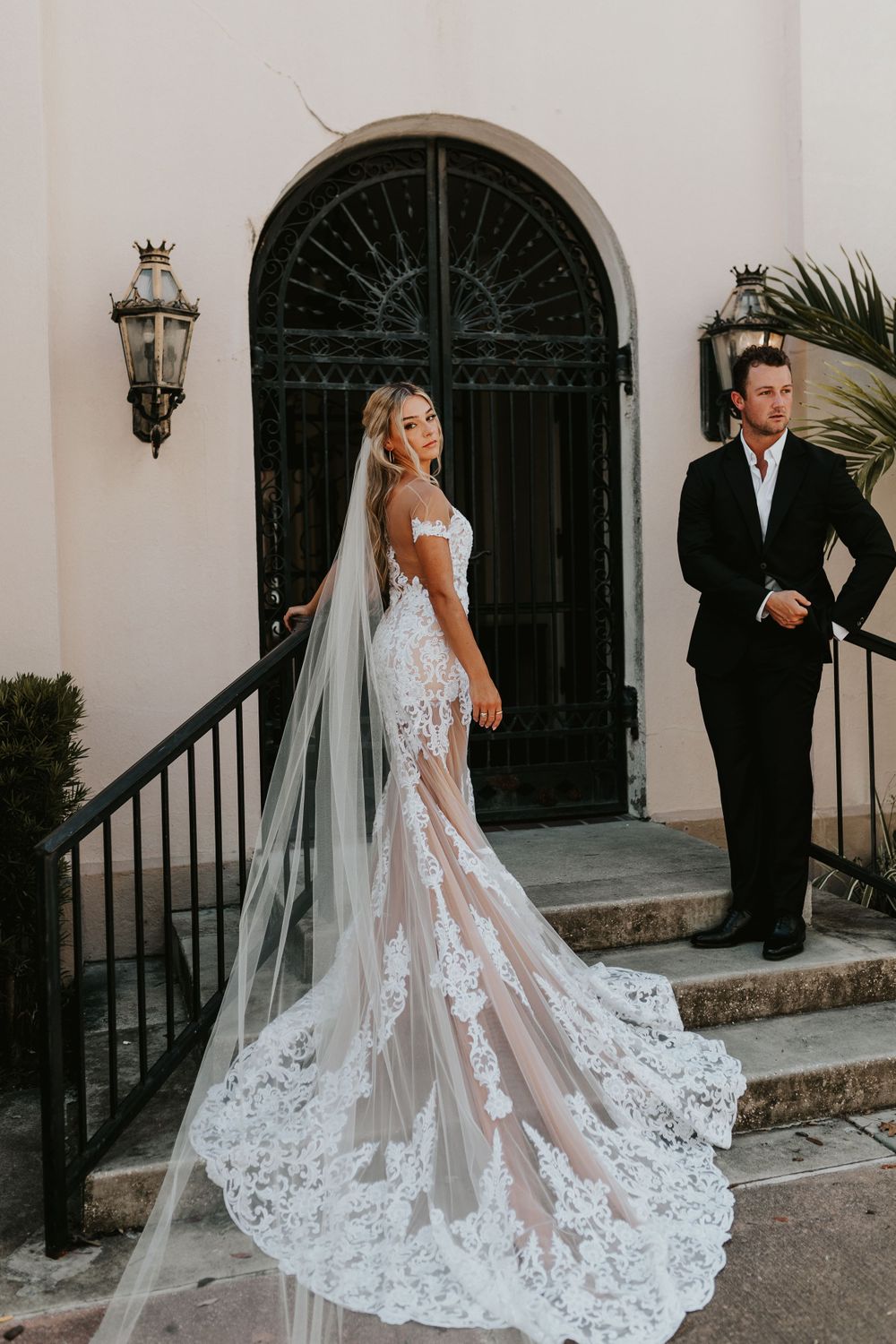 Bride in lace gown with cathedral veil poses by ornate black door while groom stands nearby.