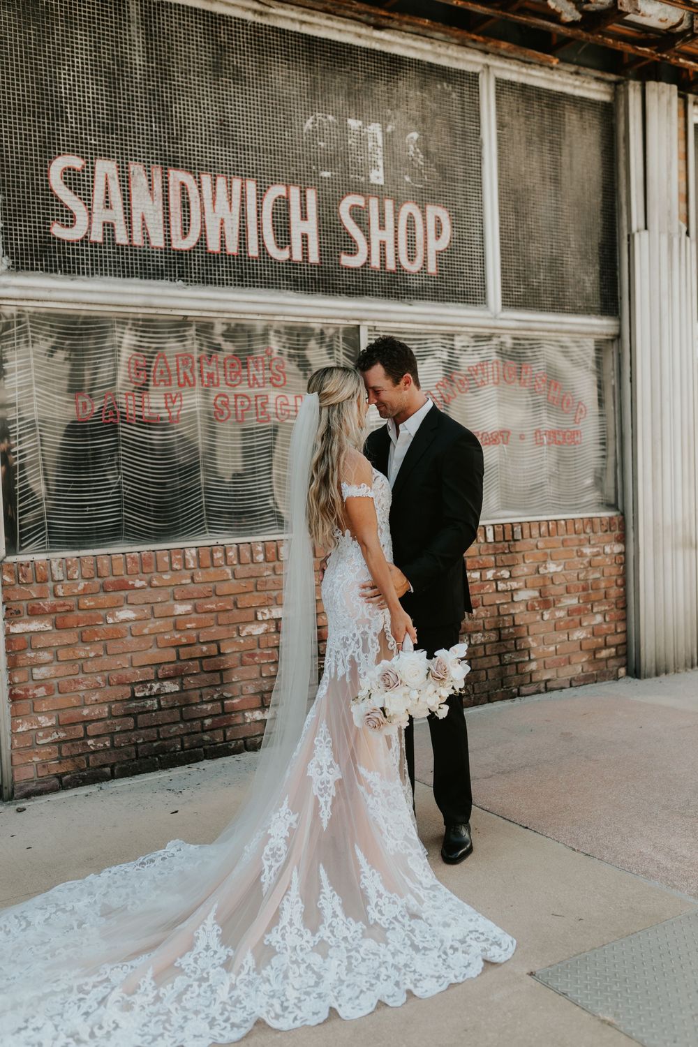 Wedding couple poses in front of rustic brick wall with vintage sandwich shop sign.