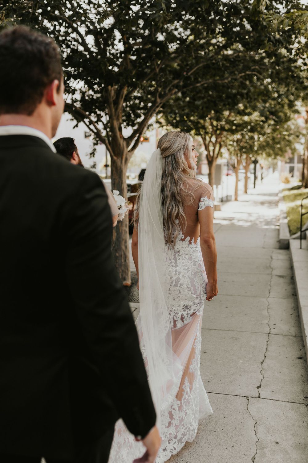 Wedding couple walking along tree-lined street with flowing veil and lace dress.