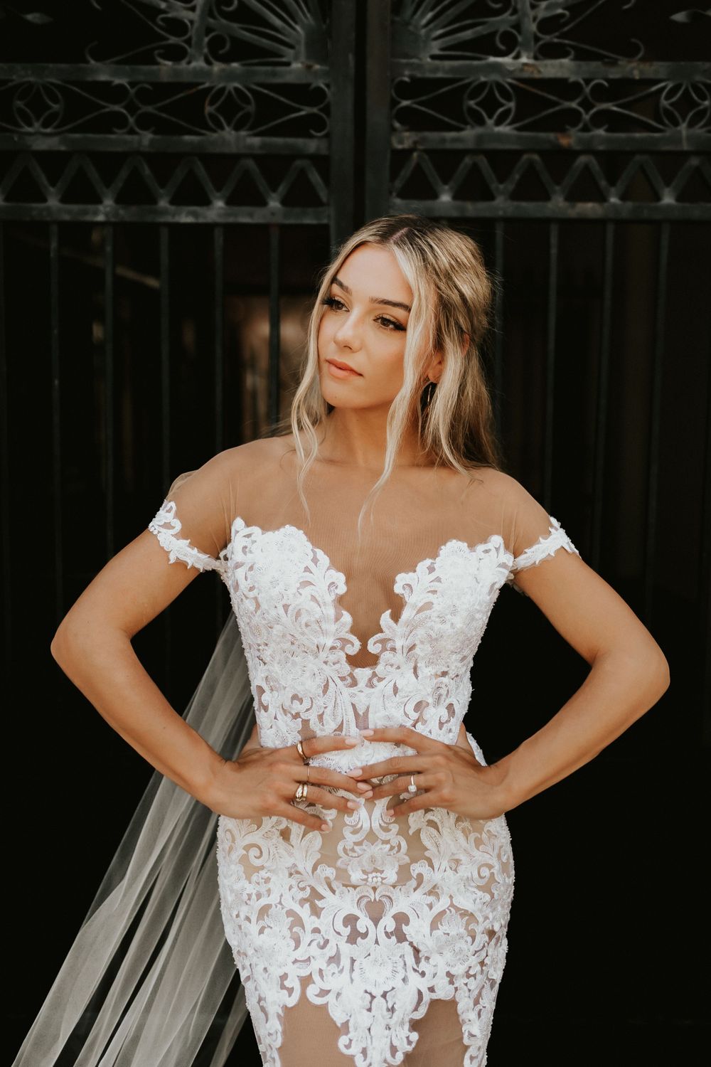 Bride in detailed lace wedding gown with sheer details poses against dark gates.