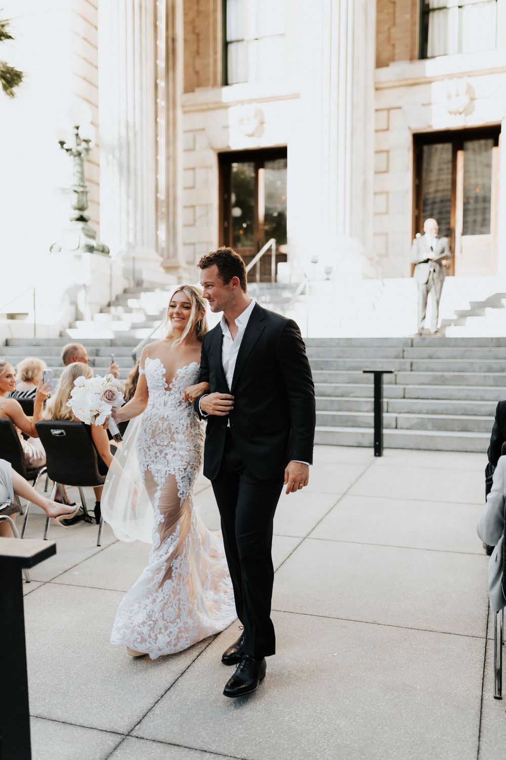 Wedding couple exits ceremony venue with classical architecture and columns.