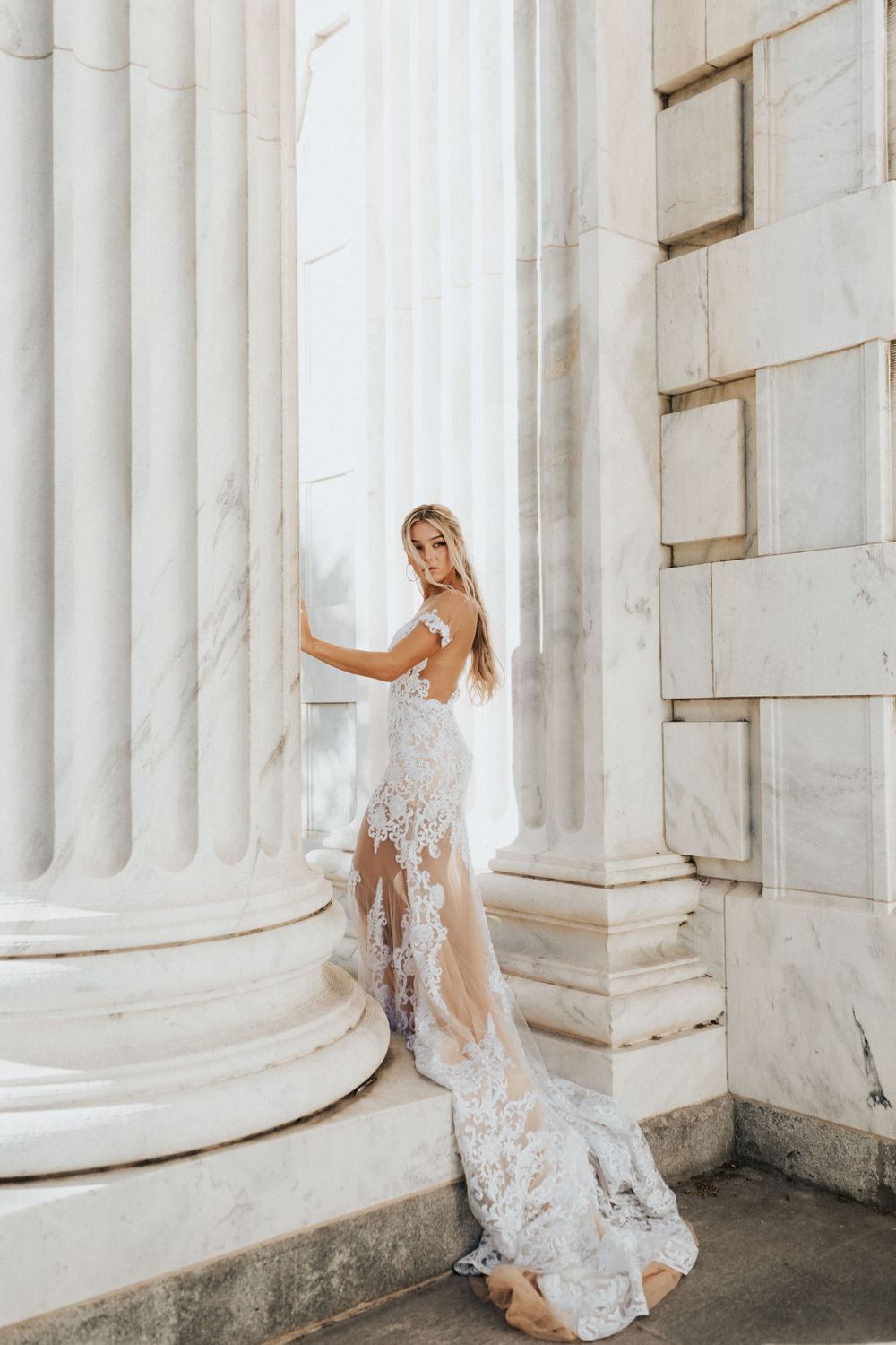 Bride in lace wedding gown poses between grand white marble columns.