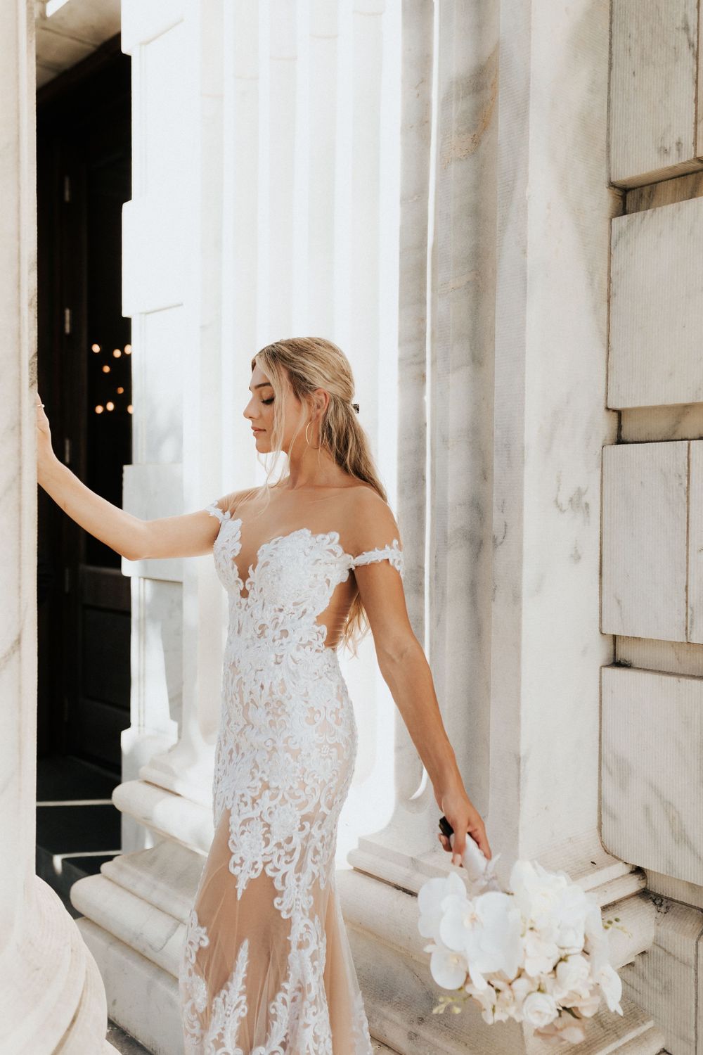Elegant bride in off-shoulder lace gown poses against white marble columns while holding a white bouquet.