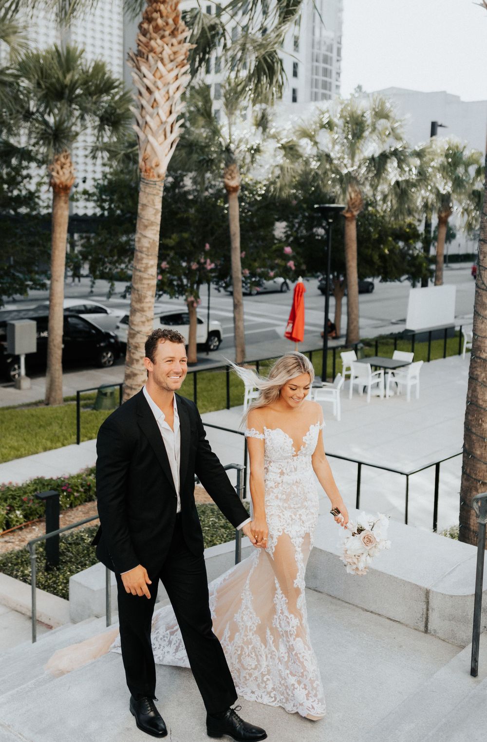 Wedding couple walks down palm-lined pathway, bride in lace gown and groom in black suit.