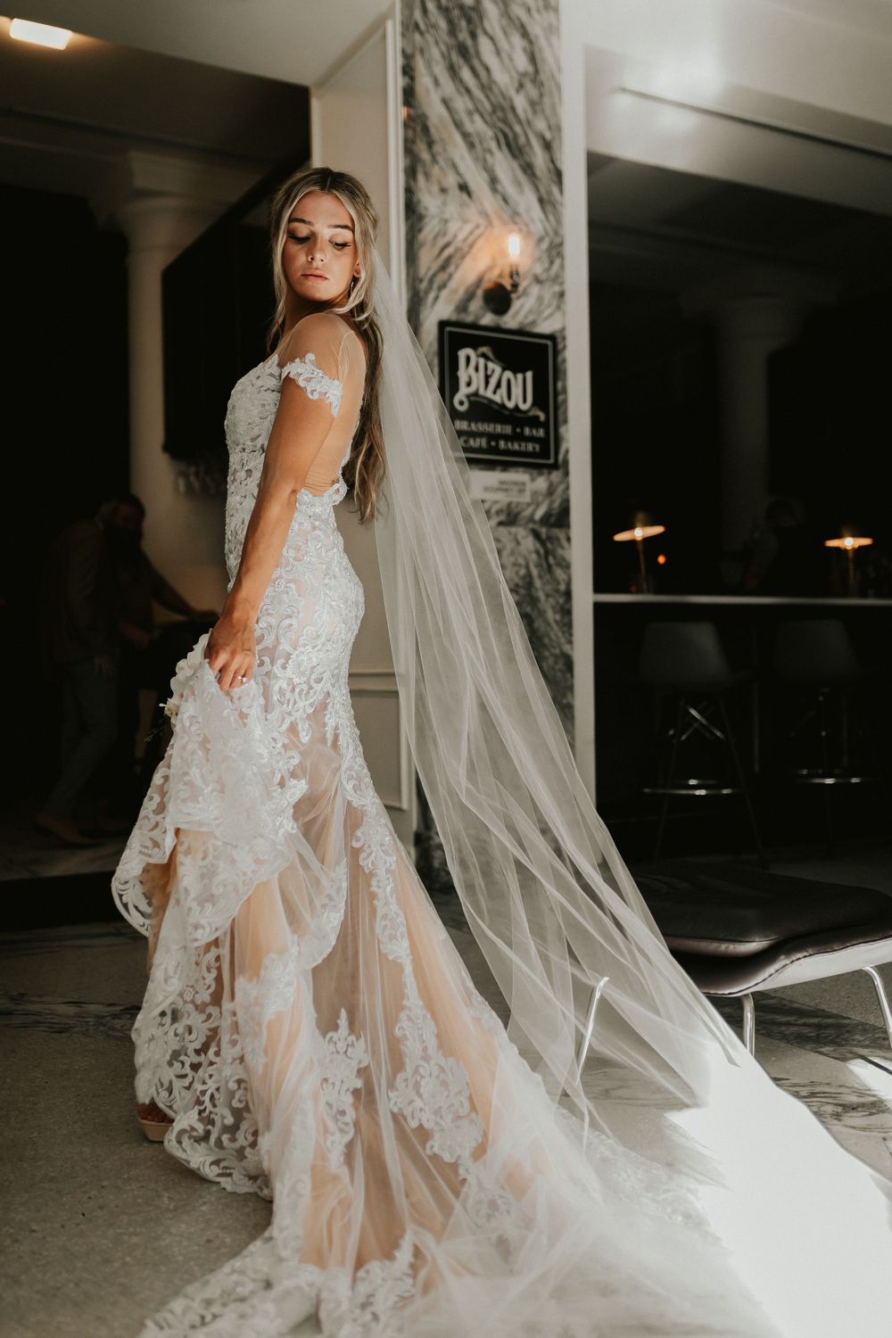 Bride in sheer lace wedding gown with long veil poses in marble hallway.