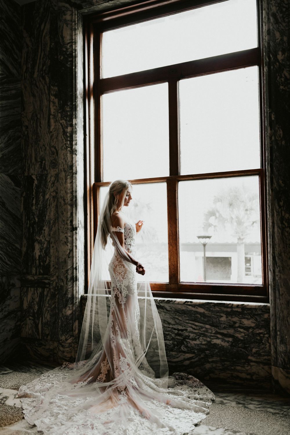 Ethereal bride in lace gown gazes through large vintage window.