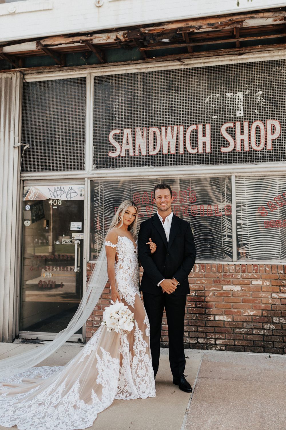 A wedding couple poses in front of a vintage sandwich shop storefront with exposed brick.