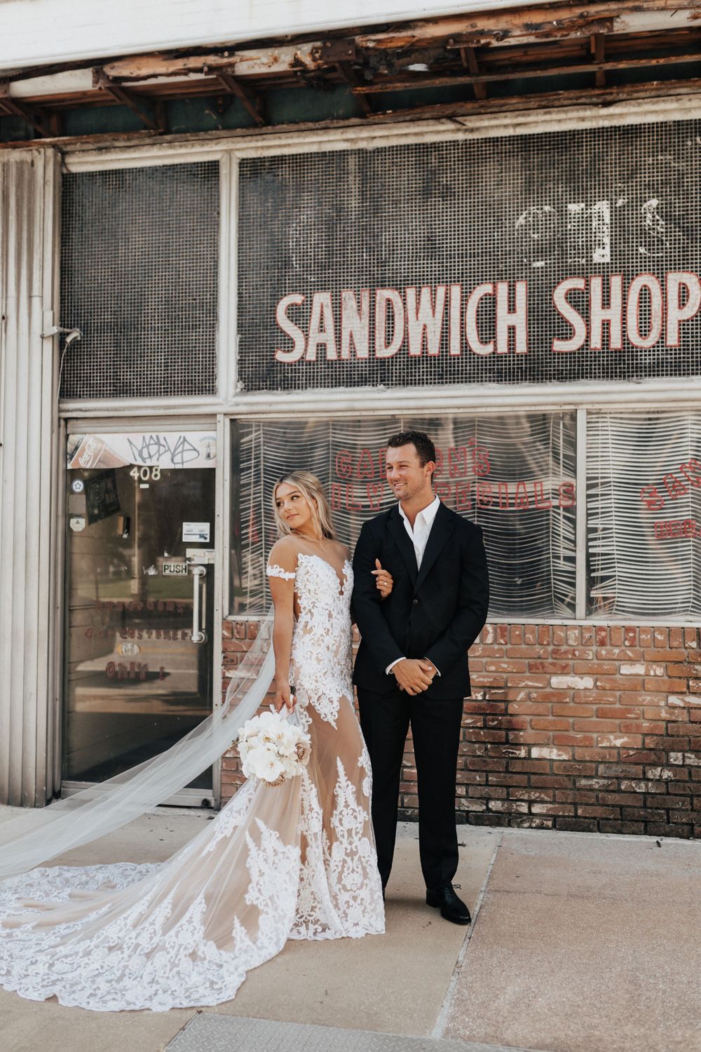 A stylish wedding couple standing in front of a vintage sandwich shop with long flowing veil.
