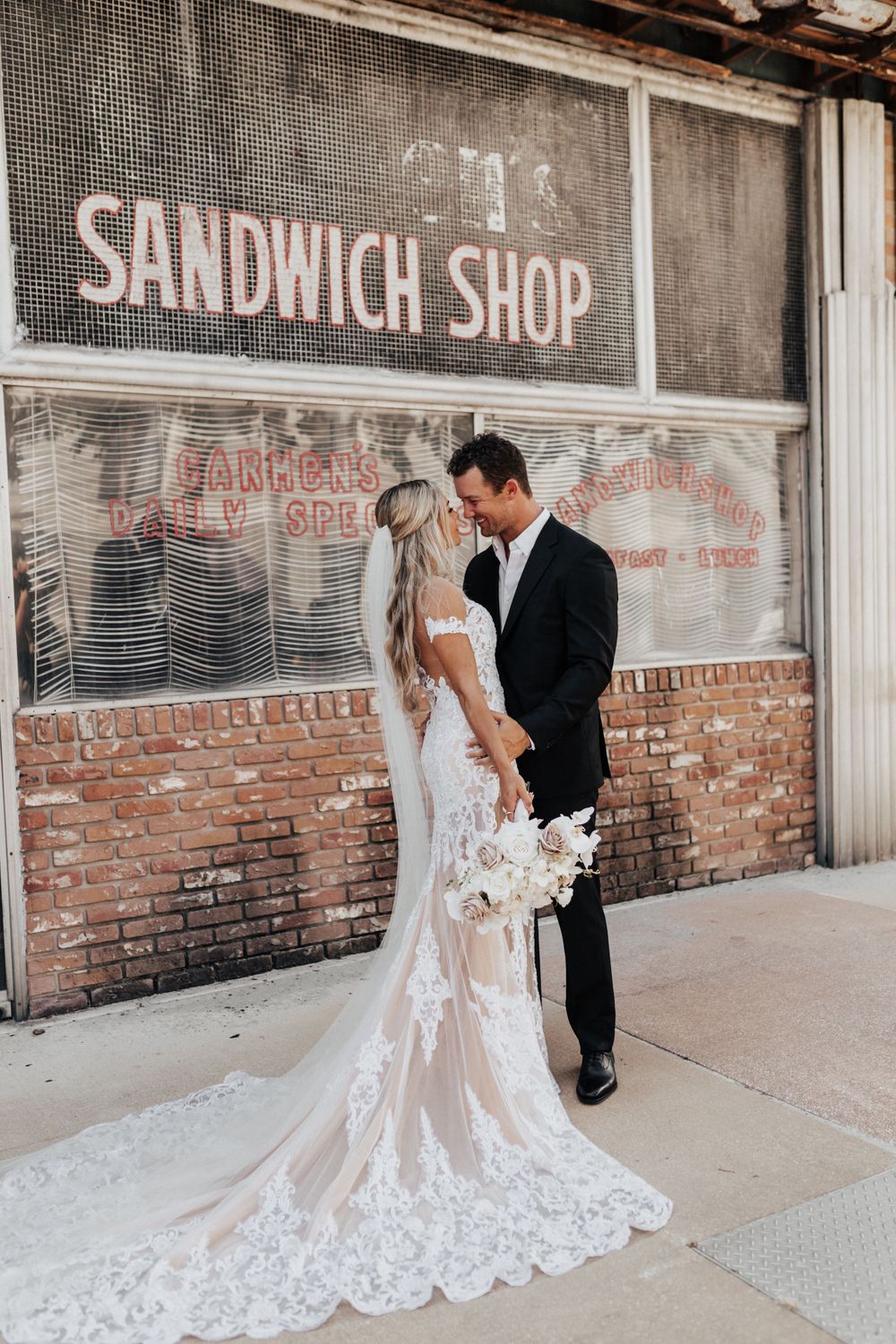A couple poses for a wedding photo outside a rustic sandwich shop with vintage signage.