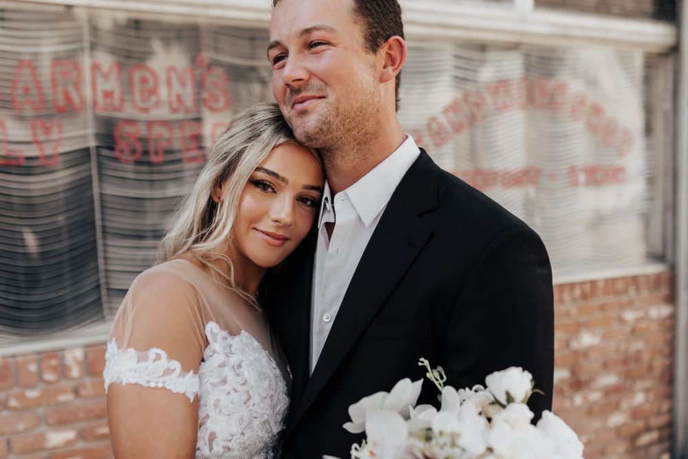 A wedding couple shares an intimate moment together while holding a white bouquet in front of a brick wall.