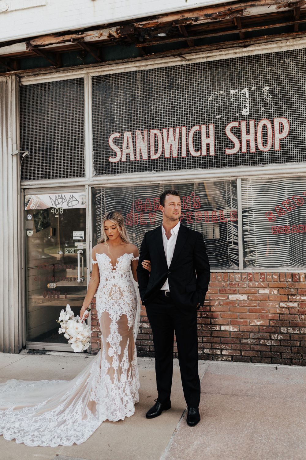 A bride in a lace gown and groom pose together outside an old sandwich shop.