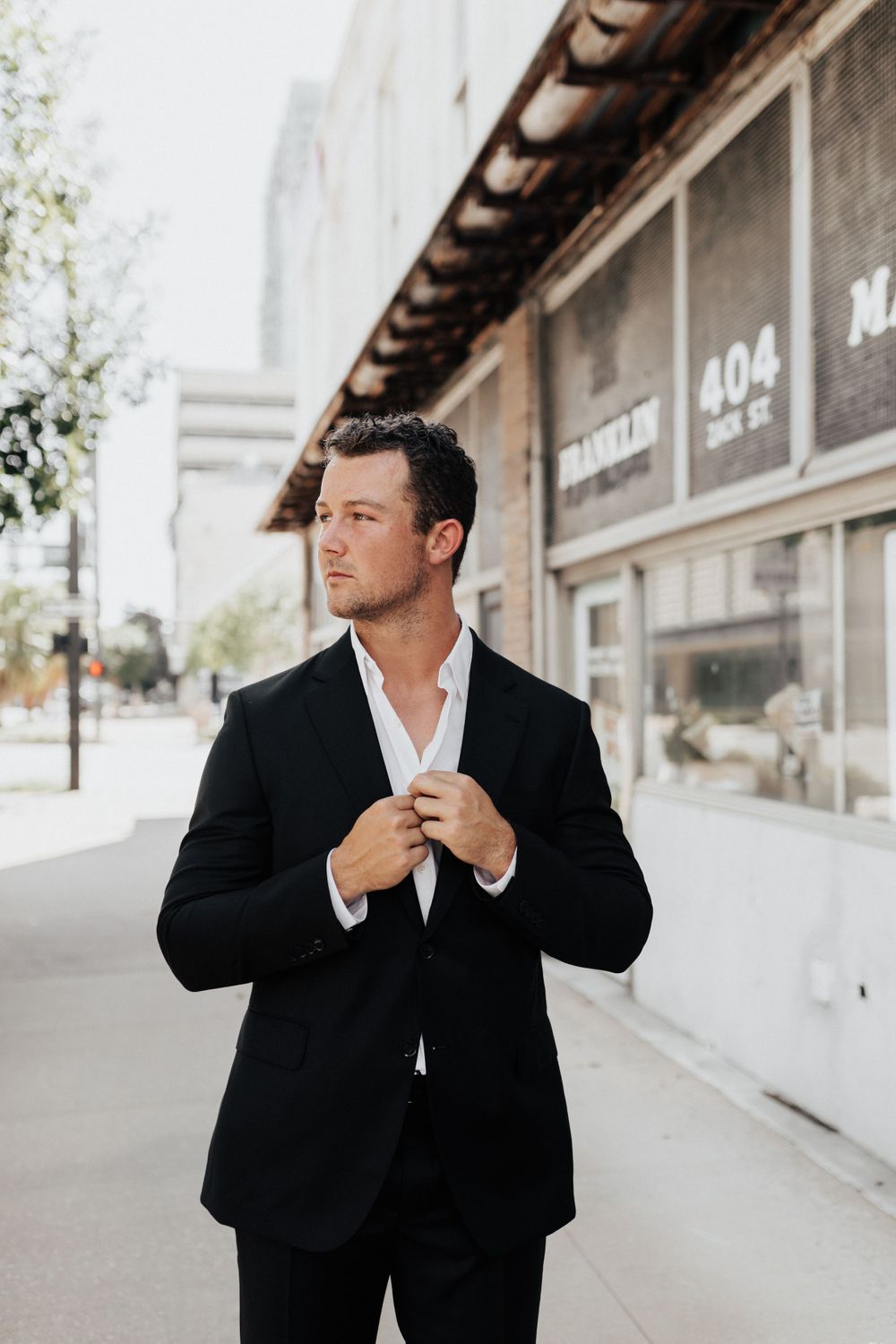 A groom in a black suit adjusts his collar while standing outside a vintage storefront.