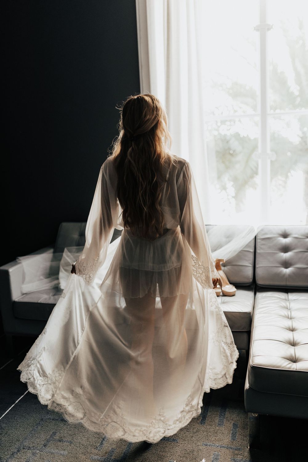 Ethereal white lace robe silhouetted against bright window with grey couch in foreground.