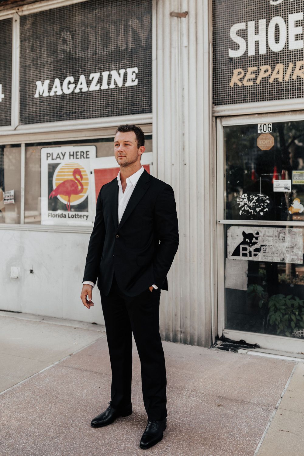 A groom in formal attire poses in front of vintage shop windows displaying magazine and shoe repair signs.