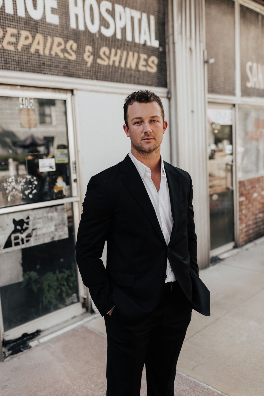 A formally dressed man stands confidently outside a vintage shoe hospital storefront.