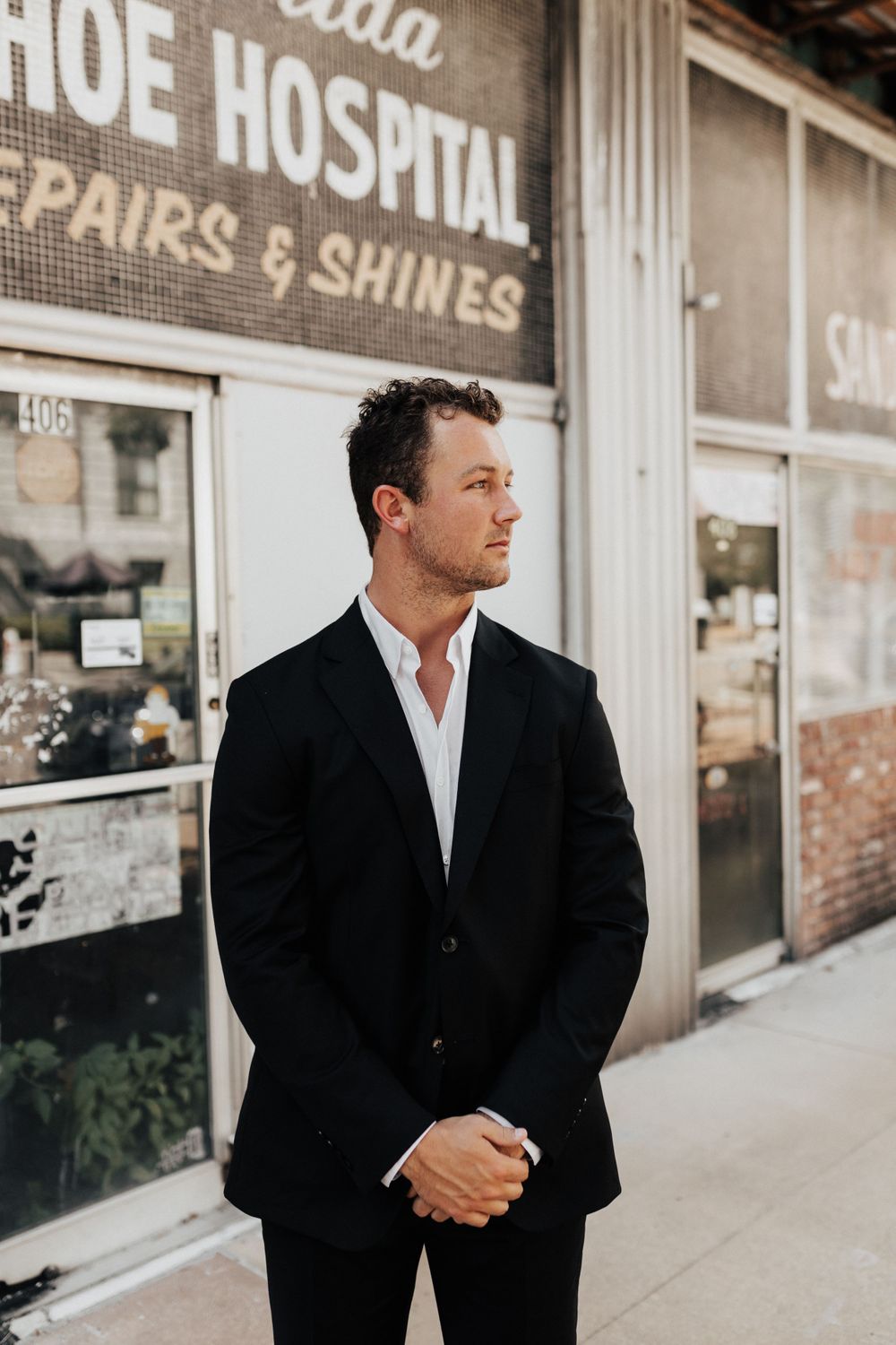 A well-dressed man in a black suit poses contemplatively outside a vintage shoe repair shop.