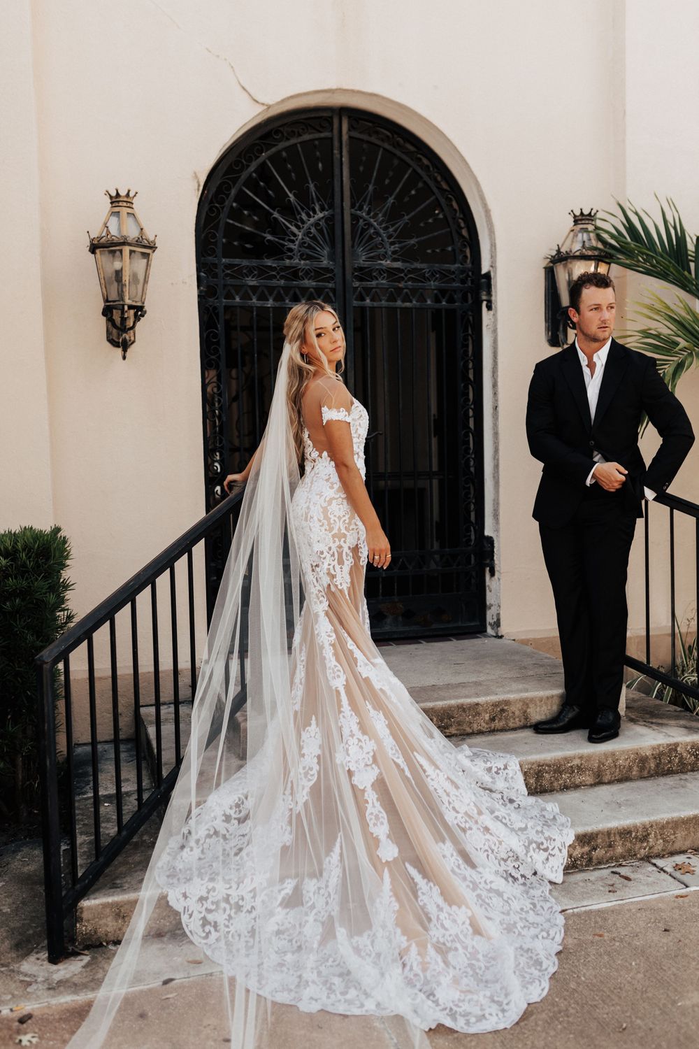 A couple poses on steps near an ornate black iron door for an elegant wedding portrait.