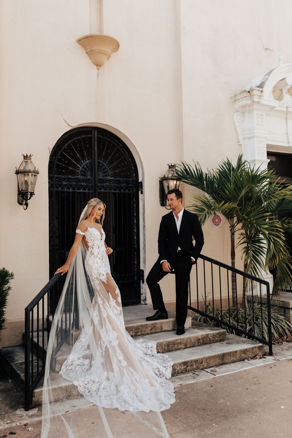 A wedding couple poses on cream-colored steps in front of an ornate black door with palm plants and lanterns.