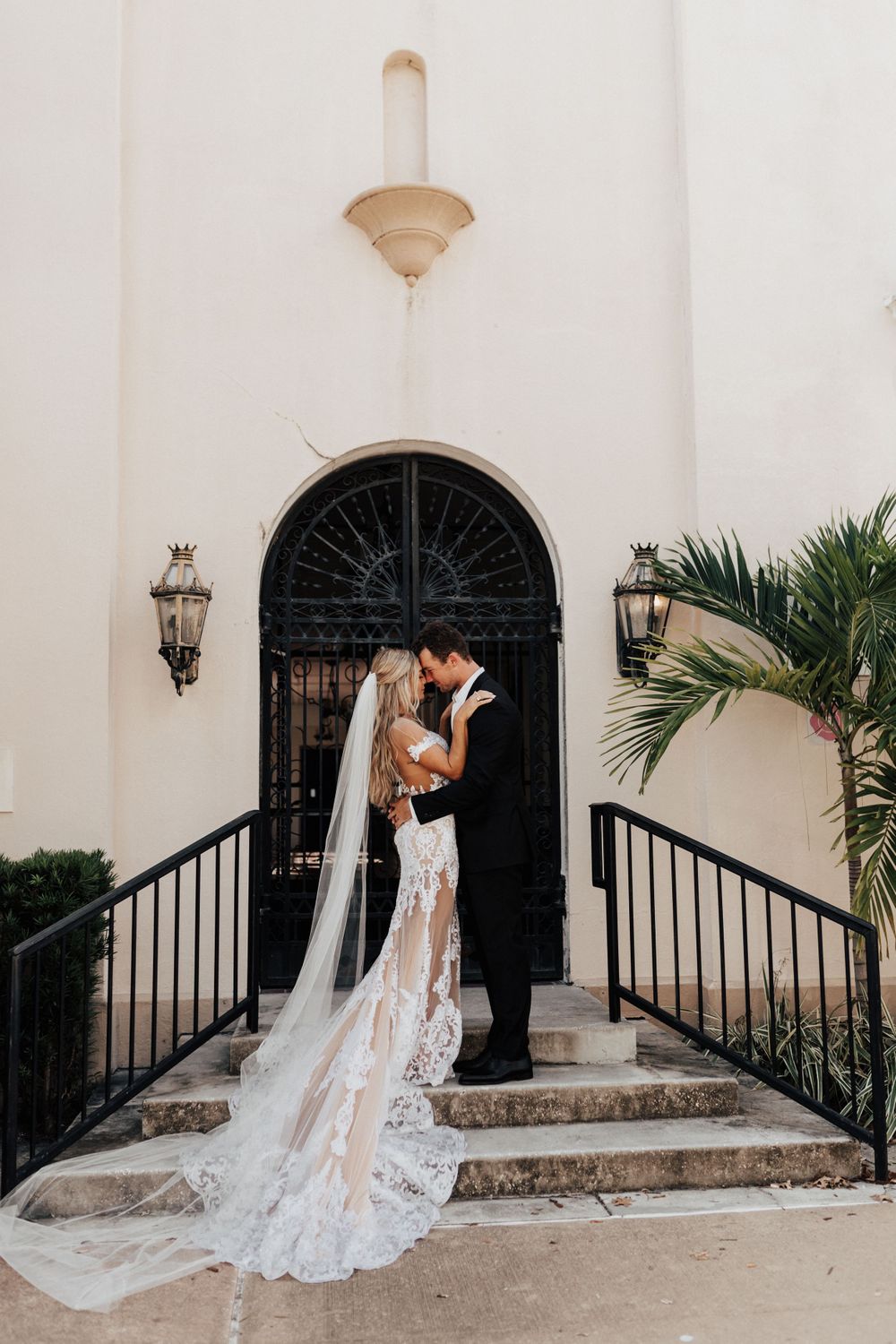 Romantic wedding moment captured on stone steps with ornate black door and flowing lace train.