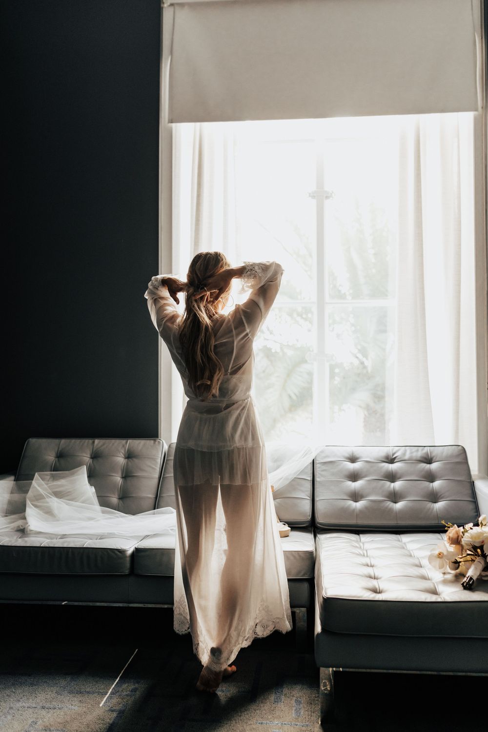 Silhouette in white robe standing at window with modern grey furniture in foreground.