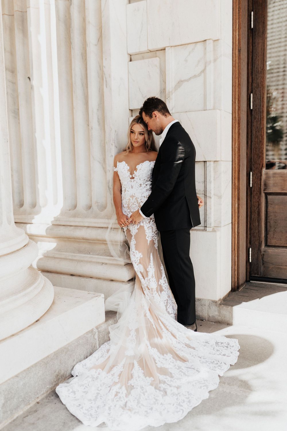 Romantic wedding portrait against white marble columns showing detailed lace gown with long train.