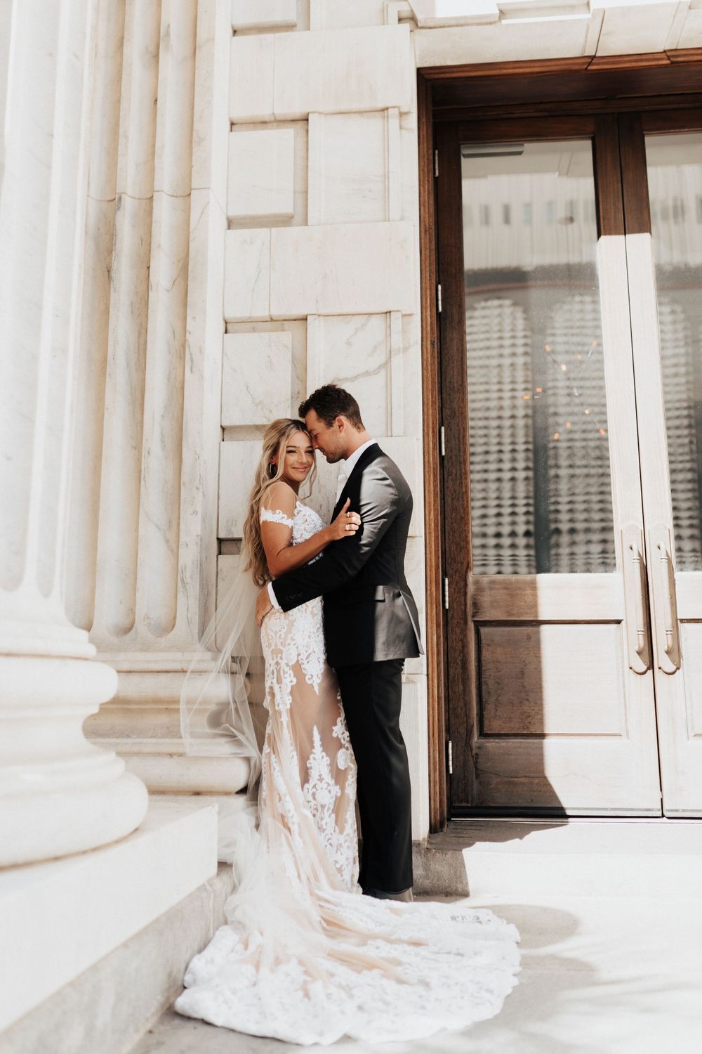 Romantic wedding portrait showing couple embracing against white marble columns and wooden doors.