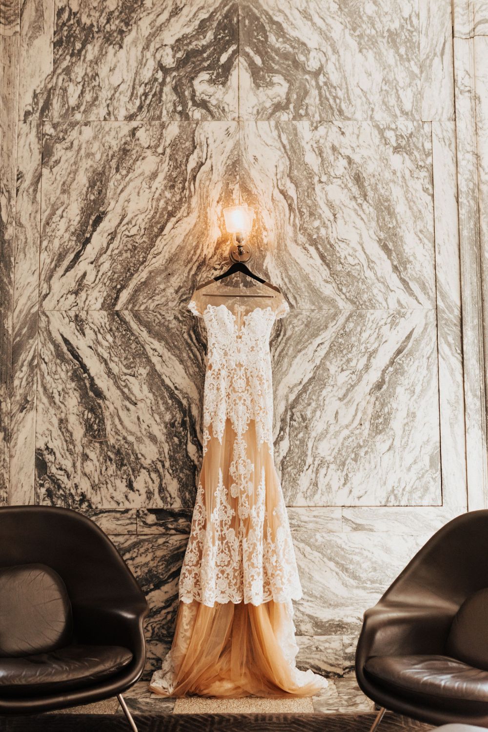 Elegant lace wedding dress displayed against dramatic marble wall with sconce lighting.