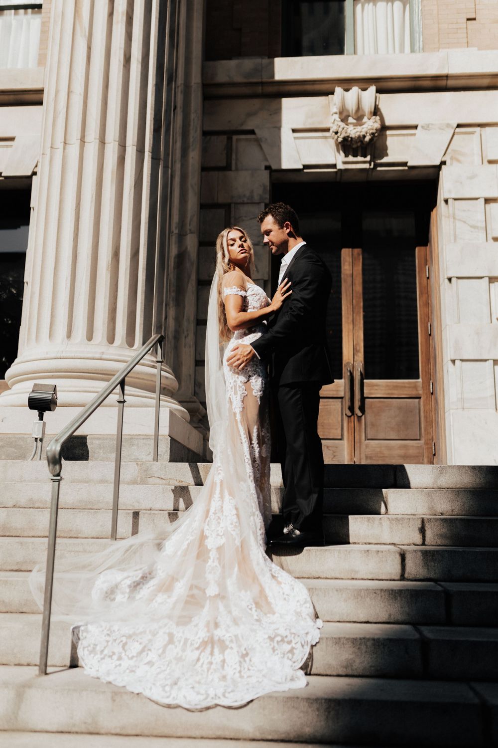 Elegant wedding couple poses on grand stone steps with classical columns and flowing lace train.